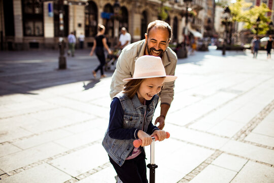 Father helping daughter ride scooter on sunny city street