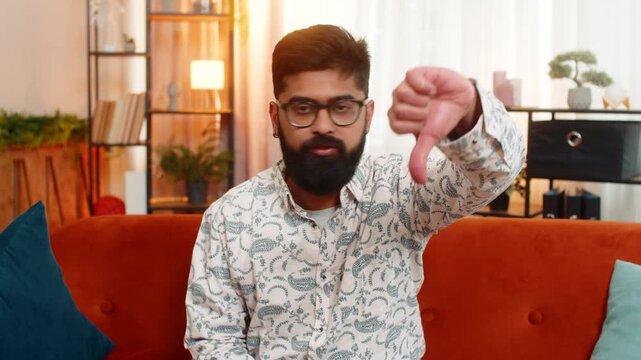 Indian man at home shows dislike with angry face, giving thumbs down gesture to express strong disapproval. Arabian guy on sofa reacts emotionally with frustration, dissatisfaction, and irritation