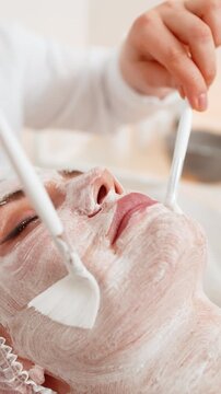 Enzyme therapy. Beautician applying moisturizing medical chemical peeling cream mask on young adult woman face chin with brushes. Girl skincare procedure on spa. Facial cosmetology treatment. Vertical