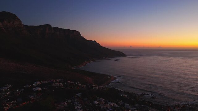 Aerial view at dusk shows Table Mountain, Twelve Apostles, and Camps Bay in Cape Town. The camera slowly surveys the bay, street lights glow, and waves lap rocky outcrops.