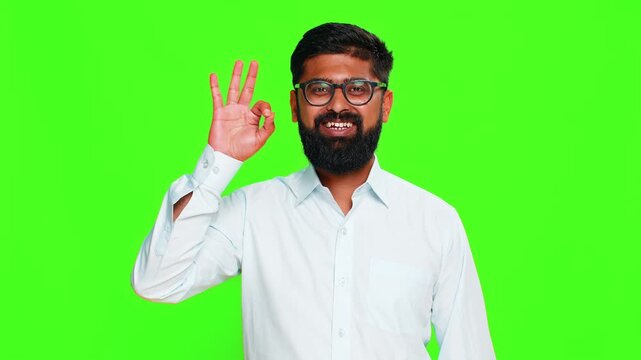 Indian man makes okay hand gesture with cheerful smile expressing agreement and positive attitude with friendly energy. Arabian guy isolated on green chroma key background showing approval and support
