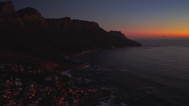 Aerial dusk over Cape Town Atlantic seaboard shows Table Mountain and the Twelve Apostles in silhouette, car lights trace the coastal road, waves break on rocky coves.