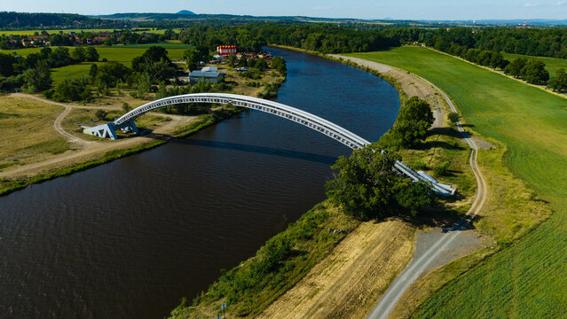Aerial view of the arched gas pipeline bridge crossing the river surrounded by lush green fields and trees in Nova Ves, Central Bohemian Region, Czechia.