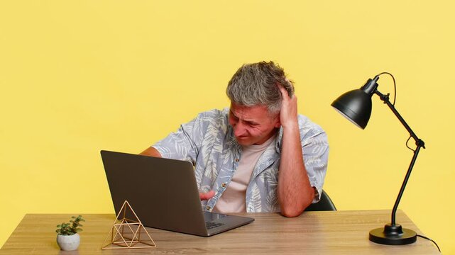 Middle-aged man holding head with both hands at table after reading laptop screen clearly frustrated and upset by error. Young guy reacts to computer failure or lost file isolated on yellow background