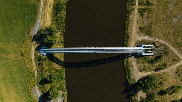 Aerial view of a gas pipeline bridge crossing a dark river surrounded by green fields and dirt paths in Nova Ves, Central Bohemian Region, Czechia.