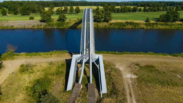 Aerial view of a gas pipeline bridge crossing a wide river surrounded by lush green fields and dense forests under a clear sky Nova Ves, Central Bohemian Region, Czechia.