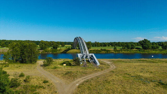 Aerial view of the arched gas pipeline bridge crossing the river surrounded by lush green meadows and trees under a clear blue sky in Nova Ves, Central Bohemian Region, Czechia.