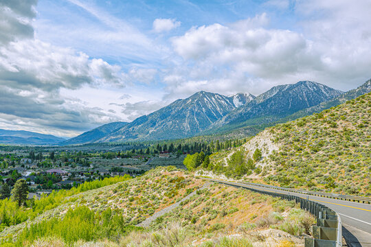 A winding mountain highway with guardrails bends down toward a small town valley near Genoa, backdropped by massive peaks
