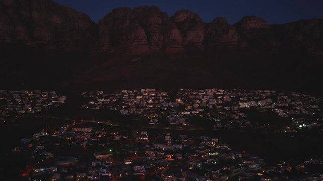 Aerial night scene in Cape Town shows the Twelve Apostles ridge as the camera sweeps across a residential slope with warm house lights and street lamps.