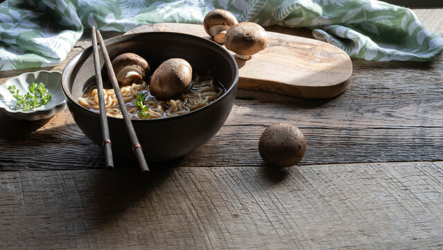 Hot noodle pho soup with whole, baby portabella mushrooms, noodles and thyme on a wooden table with green and white napkin towel and chopsticks