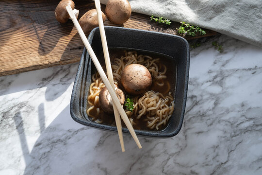 Korean style mushroom and noodles soup or pho, garnished with fresh thyme, in a black square bowl on a marbled background.