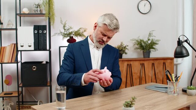 Middle-aged businessman at home office opens piggy bank shakes coins counting them feeling sad and anxious. Freelancer guy at table drafts budget forecasting debt payment hoping to prevent bankruptcy.