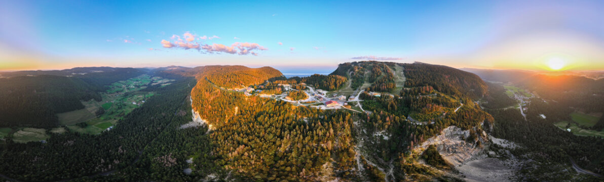 Aerial view of the Col de la Faucille mountain pass at sunset with golden light over the lush green forests and mountain ridges in Gex, Auvergne-Rhone-Alpes, France.