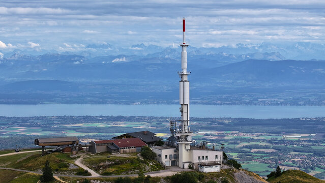Aerial view of a telecommunications tower on a mountain peak overlooking Lake Geneva and the distant Alps in Gex, Auvergne-Rhone-Alpes, France.