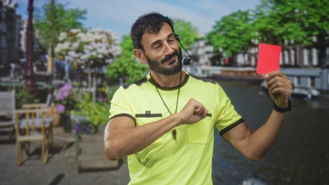 Man referee in neon yellow uniform holding a red card and pointing finger, whistle on lanyard and headset visible, beard and wristwatch, standing on street by a canal cafe; discipline authority.