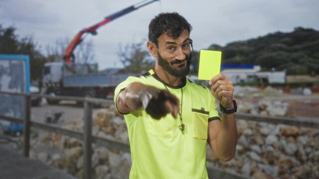 Man points finger and holds yellow card on street construction site, wearing neon shirt and whistle visible; authority warning.