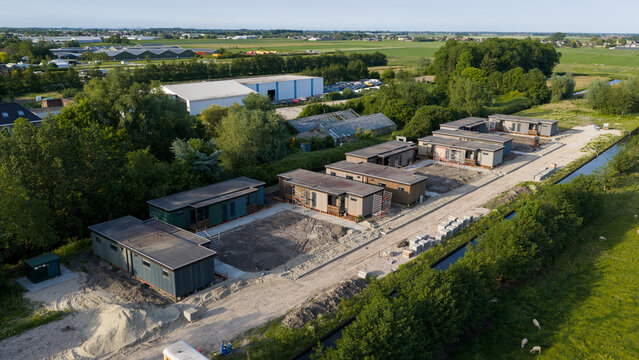 Aerial view of modular houses under construction with a dirt path, green trees, and sheep grazing in a meadow near a canal in Ter Aar, Zuid-Holland, Netherlands.