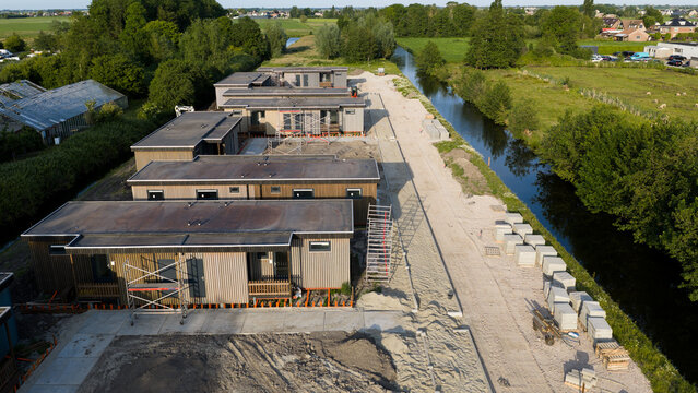 Aerial view of modern wooden modular houses under construction along a canal with scaffolding and lush greenery in Ter Aar, Zuid-Holland, Netherlands.