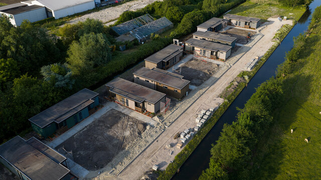 Aerial view of modular houses under construction along a canal with lush green trees and industrial buildings in Ter Aar, Zuid-Holland, Netherlands.
