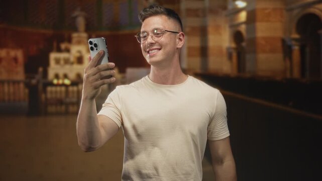 Young man holding a smartphone, smiling and waving during a video call inside a church nave with altar and pews visible; joyful greeting.