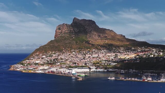 Aerial view of a Cape Town coastal town shows a rocky headland, terraced houses, and an enclosed harbor with piers and moored fishing boats in late afternoon light.