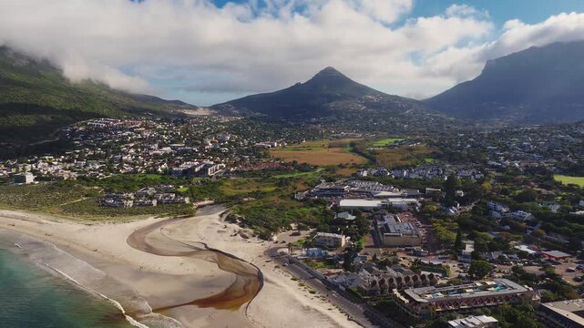 Aerial drone view of Camps Bay in Cape Town shows a crescent beach, tidal stream, Lion's Head, Table Mountain, homes and promenades, late afternoon light and clouds.