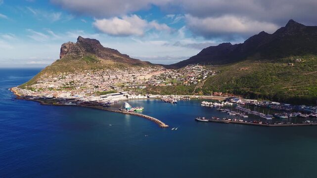 Aerial scene of Hout Bay, Cape Town, South Africa. Boats leave wakes, yachts and fishing vessels moor in the marina. Low sun warms roofs and The Sentinel.