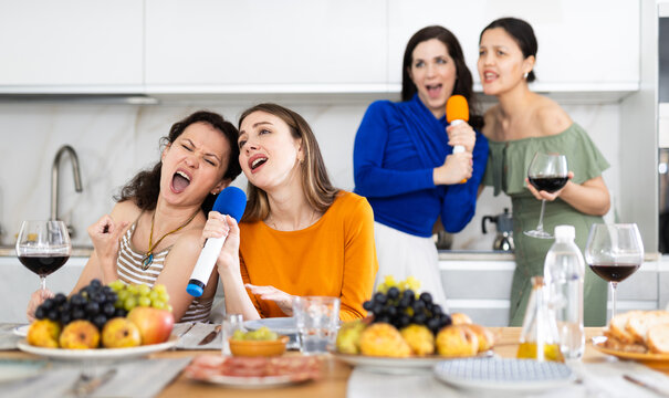 Pair of cheerful, slightly tipsy women enjoying singing with microphones at home karaoke during friendly girls get-together with wine and light snacks