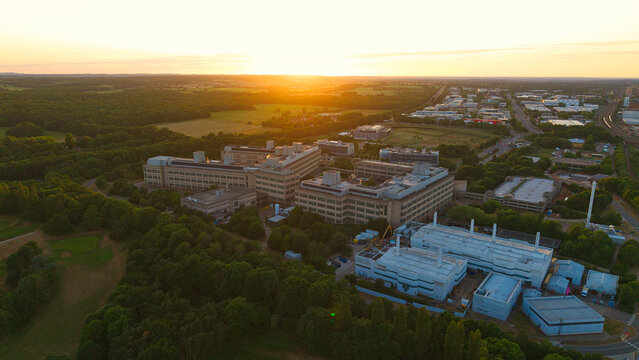 Aerial view of the GSK Stevenage research campus surrounded by dense green forests and industrial facilities during a golden sunset in Stevenage, England, United Kingdom.