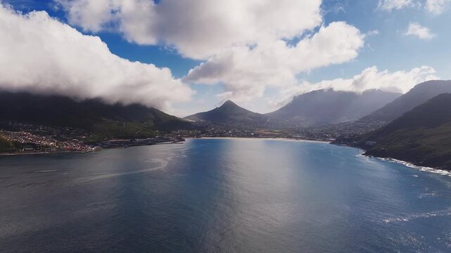Aerial view of Hout Bay in Cape Town shows Sentinel peak and Table Mountain as the camera glides over calm water, faint boat wake, rocky headlands, and moody cloud shadows.