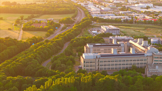 Aerial view of the GSK Stevenage research complex surrounded by lush green trees and a highway during golden hour in Stevenage, England, United Kingdom.