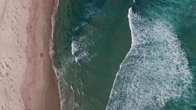 Aerial top down view shows pale sand meeting turquoise ocean near Cape Town, South Africa. Waves roll to shore, two figures walk, footprints and sandbars show motion.
