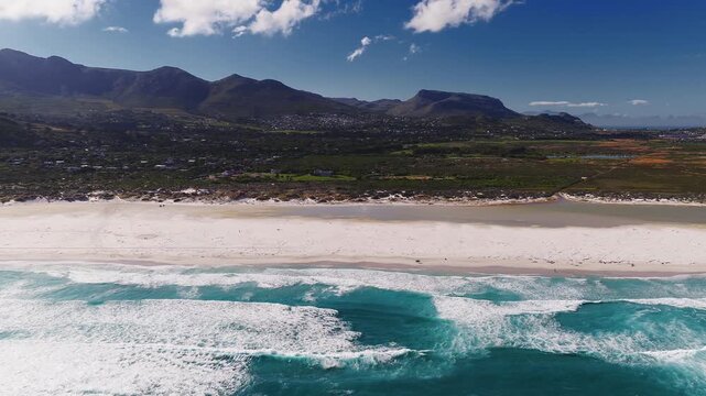 Aerial view of a long white sand beach near Cape Town, South Africa, with surf, dunes, scrub, houses, and mountains. Tiny figures and vehicles appear in bright daylight.