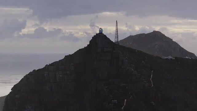 Aerial lighthouse at Cape Point near Cape Town with radio mast, Atlantic horizon, storm light, layered clouds, winding path, seabirds, rugged cliffs, pre dusk