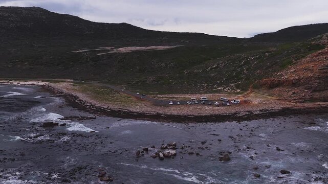 Aerial scene shows Cape of Good Hope headland near Cape Town, vans and cars at a small turnout, churning waves, offshore rocks, pebble shore, and overcast sky.
