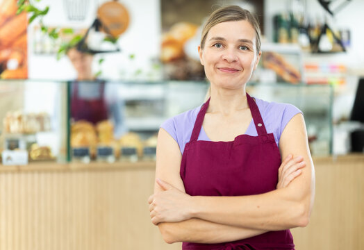 Positive young waitress wearing pinafore standing in cozy cafeteria ready to greet visitors