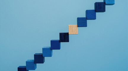 Fototapeta premium Wooden blocks form a step ladder with different blue shades. One block differs in color and texture, suggesting progress or uniqueness. Clean blue background highlights abstract concept.