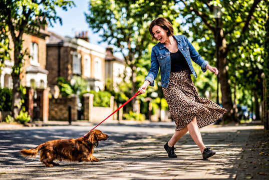 Laughing woman walking playful dachshund dog on red leash down summer street