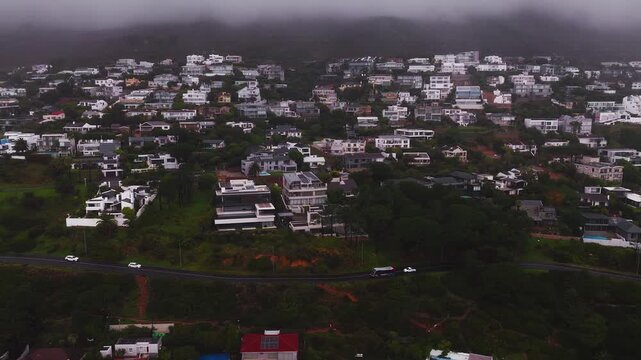 Aerial view of modern white villas with terraces and pools above a coastal road in Cape Town. Low cloud rolls over slopes, vehicles move, and muted light sets a moody tone.