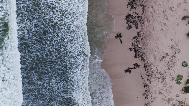 Aerial top down view of a Cape Town beach shows dark green ocean, frothy surf, pale sand, and kelp. Waves break and recede in overcast daylight, forming lace patterns.