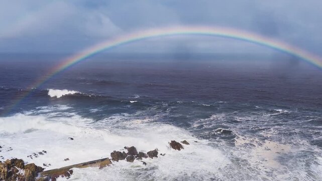 High angle aerial view shows Cape of Good Hope surf, jagged rocks, and a low concrete jetty remnant as waves crash and a vivid rainbow arcs above under storm clouds.