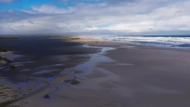 Aerial panoramic view of Western Cape beach near Cape Town with lagoon meeting the sea, ebb channels and pools in wet sand, rows of waves, and storm clouds with sunlight.