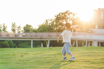 Obraz premium A young Asian boy plays with a toy airplane on a lush green lawn during golden hour sunset, with a pedestrian bridge and trees in the background.