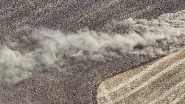 top mid-range view of farmland where a tractor is driving to clean crops and prepare earth, create a dust clouds, and divides land diagonally into two distinguish sections.
