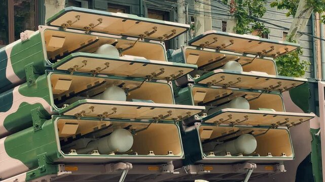 A detailed close look at a drone inside its launcher transitioning to a wide shot of multiple camouflage military trucks parked on a city street during an exhibition.
