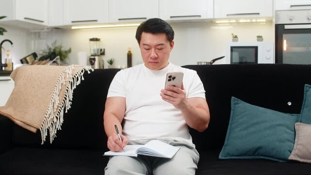 Asian man at home checks budget on smartphone calculator writes plans in notebook reviews costs. Chinese guy on sofa focuses on numbers sets goal for week saves plan to app and feels control and calm.
