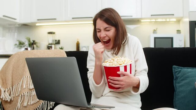 Young woman at home watches football on laptop jumps with joy at goal claps shows like eats popcorn. Caucasian girl on sofa chants for team checks score waves and enjoys stream highlight live in app