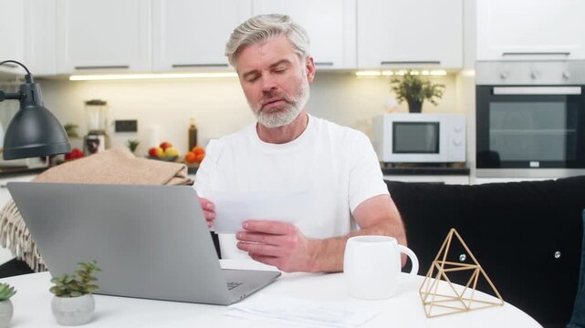 Middle-aged man at home updates budget on laptop adds expenses income lines and checks each bill carefully. Mature guy groups receipts sets priority list tracks progress adjusts categories keeps order
