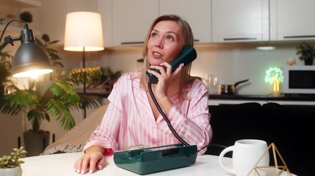 Adult woman at night home works as hotline operator uses retro corded phone listens with care. Caucasian girl at table on sofa at evening asks clarifying questions notes details finds solution fast.