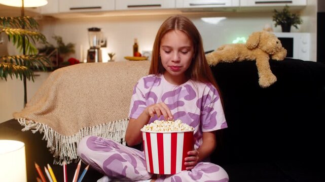 Teenager girl at night home watches movie takes popcorn from bowl laughs loud fun comedy scene. Caucasian child kid on sofa at evening chews snack smiles wipes tears of laugh enjoys leisure time joy.